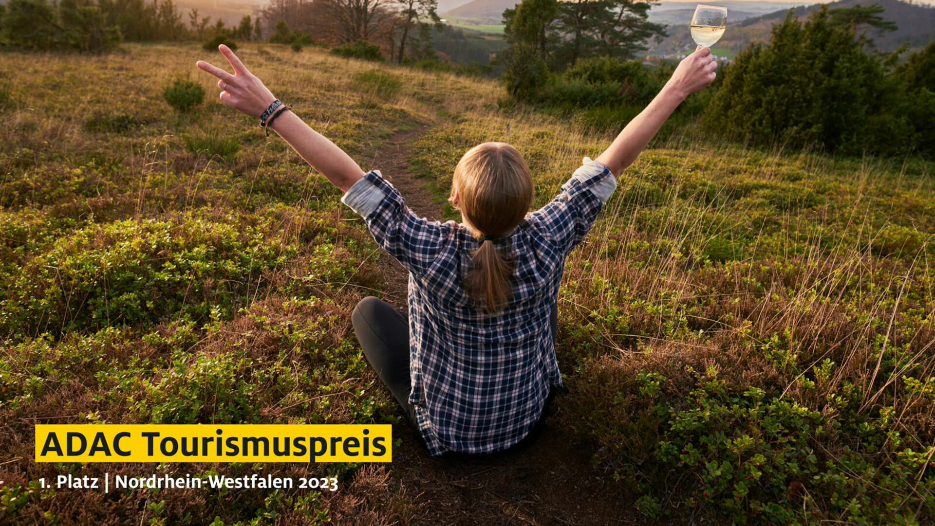 Frau sitzt auf Wiese, Blick auf Landschaft, Hände in die Luft gerichtet